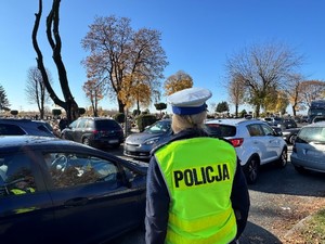 Zdjęcie przedstawia umundurowaną policjantkę stojącą w okolicach cmentarza.