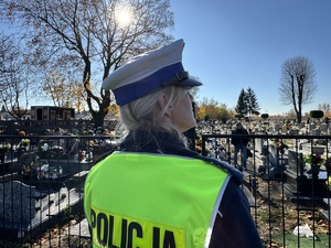 Zdjęcie przedstawia umundurowaną policjantkę stojącą w okolicach cmentarza.