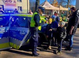 Zdjęcie przedstawia umundurowaną policjantkę z grupą dzieci w rejonie cmentarza. W tle oznakowany radiowóz policyjny.