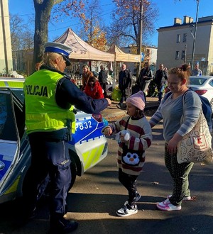 Zdjęcie przedstawia umundurowaną policjantkę wręczającą dziecku element odblaskowy. W tle oznakowany radiowóz policyjny.