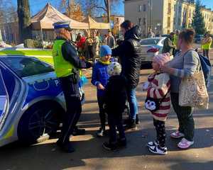 Zdjęcie przedstawia umundurowaną policjantkę z grupą ludzi w rejonie cmentarza. W tle oznakowany radiowóz policyjny.