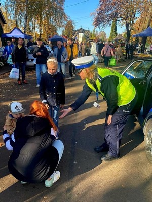 Zdjęcie przedstawia umundurowaną policjantkę z grupą ludzi w rejonie cmentarza. W tle oznakowany radiowóz policyjny.