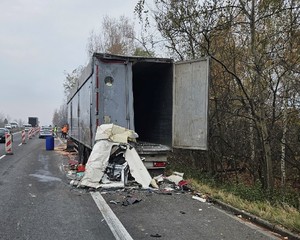 Zdjęcie przedstawia uszkodzony pojazd ciężarowy stojący na poboczu jezdni. W tle oznakowany radiowóz policyjny oraz umundurowany policjant.