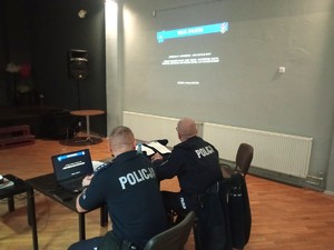 Zdjęcie przedstawia umundurowanych policjantów siedzących przy stole. W tle prezentacja multimedialna.