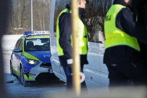 Zdjęcie przedstawia dwóch, umundurowanych policjantów w kamizelce odblaskowej oraz oznakowany radiowóz.