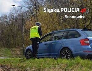 Zdjęcie przedstawia umundurowanego policjanta stojącego przy oznakowanym radiowozie podczas czynności służbowych.