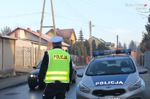 Zdjęcie przedstawia umundurowanego policjanta z wydziału ruchu drogowego podczas czynności służbowych. Obok oznakowany radiowóz.