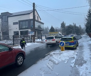 Zdjęcie przedstawia umundurowanego policjanta wydziału ruchu drogowego podczas akcji. W tle samochody i oznakowany radiowóz.