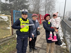 Zdjęcie przedstawia umundurowanego policjanta, dwie kobiety oraz dziewczynkę. Policjant trzyma w ręku element odblaskowy.