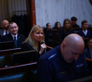 Zdjęcie przedstawia umundurowanego policjanta oraz uczestników debaty w sali.