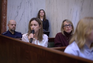 Zdjęcie przedstawia umundurowanego policjanta, przemawiająca kobietę oraz uczestników debaty w sali.