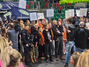 Zdjęcie przedstawia umundurowanego policjanta z psem służbowym stojącego z grupą ludzi na scenie.