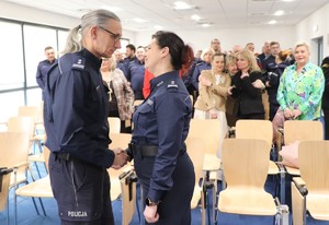 Zdjęcie przedstawia umundurowaną policjantkę i policjanta. W tle zgromadzeni uczestnicy uroczystości.