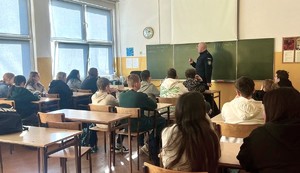 Zdjęcie przedstawia umundurowanego policjanta przemawiającego do uczniów w sali lekcyjnej. W tle tablica szkolna.