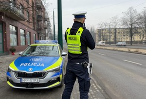 Zdjęcie przedstawia mundurowego z wydziału ruchu drogowego kontrolującego prędkość. W tle oznakowany radiowóz policyjny,