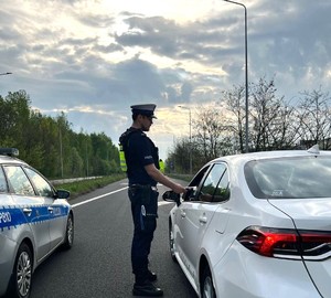Zdjęcie przedstawia umundurowanego policjanta wydziału ruchu drogowego podczas kontroli stanu trzeźwości kierowcy. W tle radiowóz.