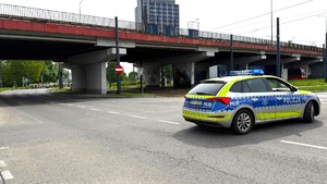 Zdjęcie przedstawia oznakowany radiowóz policyjny. W tle uczestnicy wydarzenia na rowerach w kamizelkach odblaskowych.