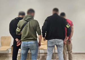 Zdjęcie przedstawia dwóch, nieumundurowanych policjantów z opaskami z napisem &quot;Policja&quot; oraz dwóch zatrzymanych mężczyzn z kajdankami spiętymi z tyłu.