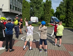 Zdjęcie przedstawia umundurowanego policjanta wydziału ruchu drogowego, oznakowany radiowóz służbowy oraz uczniów pozujących do zdjęcia.