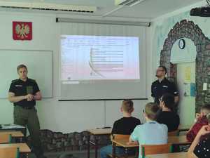 Zdjęcie przedstawia umundurowanego policjanta i funkcjonariusza Straży Granicznej w sali podczas prowadzonych zajęć. W tle prezentacja multimedialna.