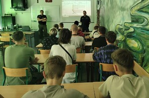 Zdjęcie przedstawia umundurowanego policjanta i funkcjonariusza Straży Granicznej w sali podczas prowadzonych zajęć. W tle prezentacja multimedialna.