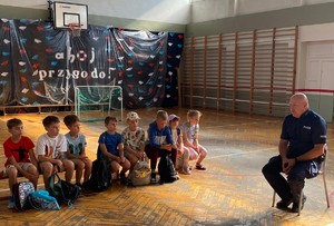 Zdjęcie przedstawia umundurowanego policjanta przeprowadzającego prelekcję z uczestnikami półkolonii.