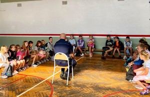 Zdjęcie przedstawia umundurowanego policjanta przeprowadzającego prelekcję z uczestnikami półkolonii.