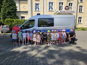 Zdjęcie przedstawia grupę przedszkolaków pozujących do zdjęcia z policjantami i oznakowanym radiowozem.