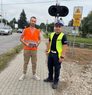 Zdjęcie przedstawia umundurowanego policjanta oraz pracownika przy przejeździe kolejowo-drogowym.