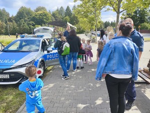 Zdjęcie przedstawia umundurowanego policjanta, oznakowany radiowóz oraz grupę ludzi.