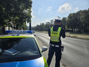 Zdjęcie przedstawia umundurowanego policjanta oraz oznakowany radiowóz służbowy podczas działań.