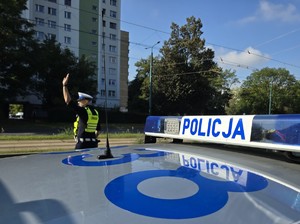 Zdjęcie przedstawia umundurowanego policjanta oraz oznakowany radiowóz służbowy podczas działań.