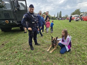 Zdjęcie przedstawia umundurowanego policjanta, psa służbowego oraz dzieci podczas pikniku.