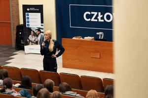 Zdjęcie przedstawia umundurowaną policjantkę z mikrofonem w sali.