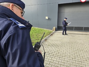 Zdjęcie przedstawia umundurowanego policjanta. W tle plecak znajdujący się przy garażu budynku komendy.