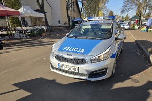 Zdjęcie przedstawia oznakowany radiowóz policyjny na tle cmentarza.