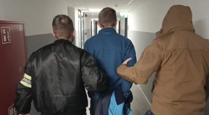 Zdjęcie przedstawia dwóch, nieumundurowanych policjantów prowadzących przez korytarz zatrzymanego mężczyznę z kajdankami zapiętymi z tyłu.