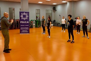 Zdjęcie przedstawia instruktora policyjnego prowadzącego zajęcia dla uczestniczek kursu w sali.