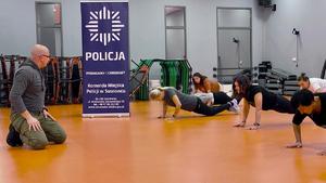 Zdjęcie przedstawia instruktora policyjnego prowadzącego zajęcia dla uczestniczek kursu w sali.