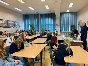 Zdjęcie przedstawia umundurowanego policjanta przemawiającego do grupy uczniów w sali lekcyjnej.