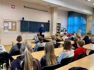 Zdjęcie przedstawia umundurowanego policjanta przemawiającego do grupy uczniów w sali lekcyjnej.