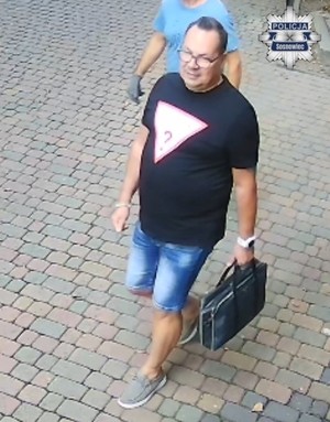 Zdjęcie przedstawia mężczyznę w okularach, ubranego w czarną koszulkę z logo oraz spodenkach jeansowych.