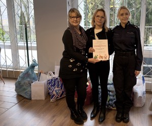 Zdjęcie przedstawia pracownicę i umundurowaną policjantkę z KMP w Sosnowcu wraz z dyrektor ośrodka. W tle zebrane przedmioty dla wychowanków.