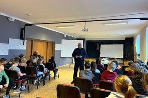 Zdjęcie przedstawia umundurowanego policjanta podczas prelekcji z uczniami w sali.