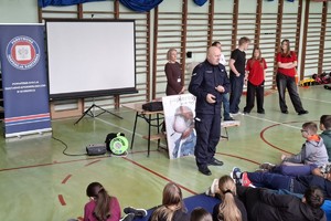 Zdjęcie przedstawia umundurowanego policjanta przemawiającego do uczniów w sali gimnastycznej.