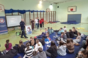 Zdjęcie przedstawia umundurowanego policjanta przemawiającego do uczniów w sali gimnastycznej.