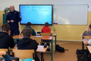 Zdjęcie przedstawia umundurowanego policjanta w sali lekcyjnej z uczniami.