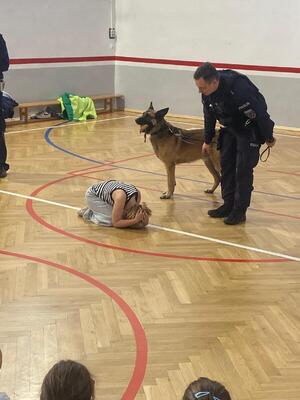 Zdjęcie przedstawia policjanta wraz z psem służbowym podczas wizyty w szkole.
