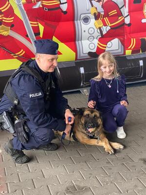 Zdjęcie przedstawia umundurowanego policjanta wraz z psem służbowym oraz dziewczynkę