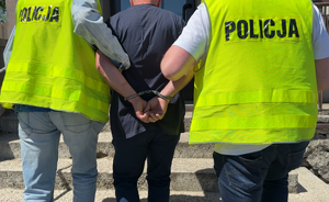 Na zdjęciu widać dwóch nieumundurowanych policjantów w kamizelkach odblaskowych, którzy prowadzą zatrzymanego do budynku komisariatu.